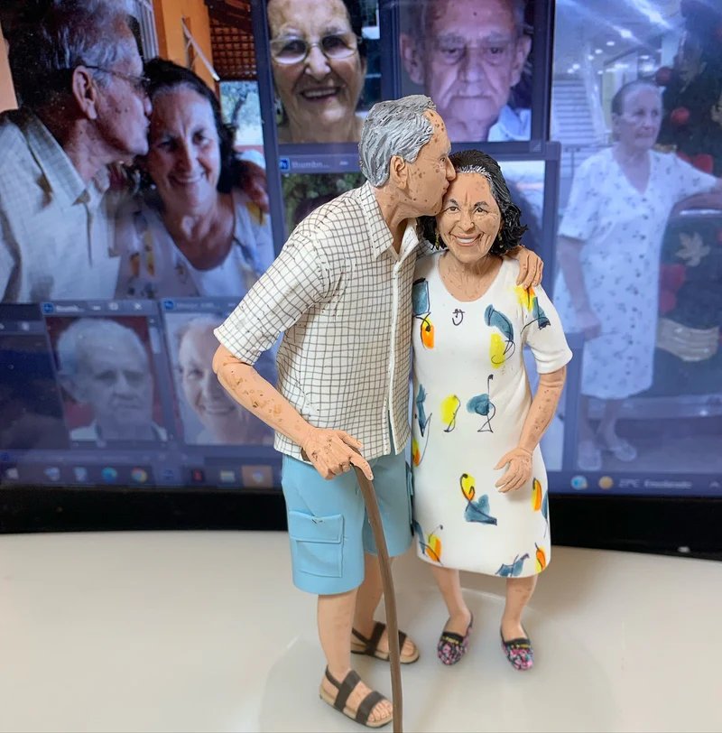 Miniatura esculpida de casal de idosos com o homem beijando a mulher na testa, com a foto original ao fundo